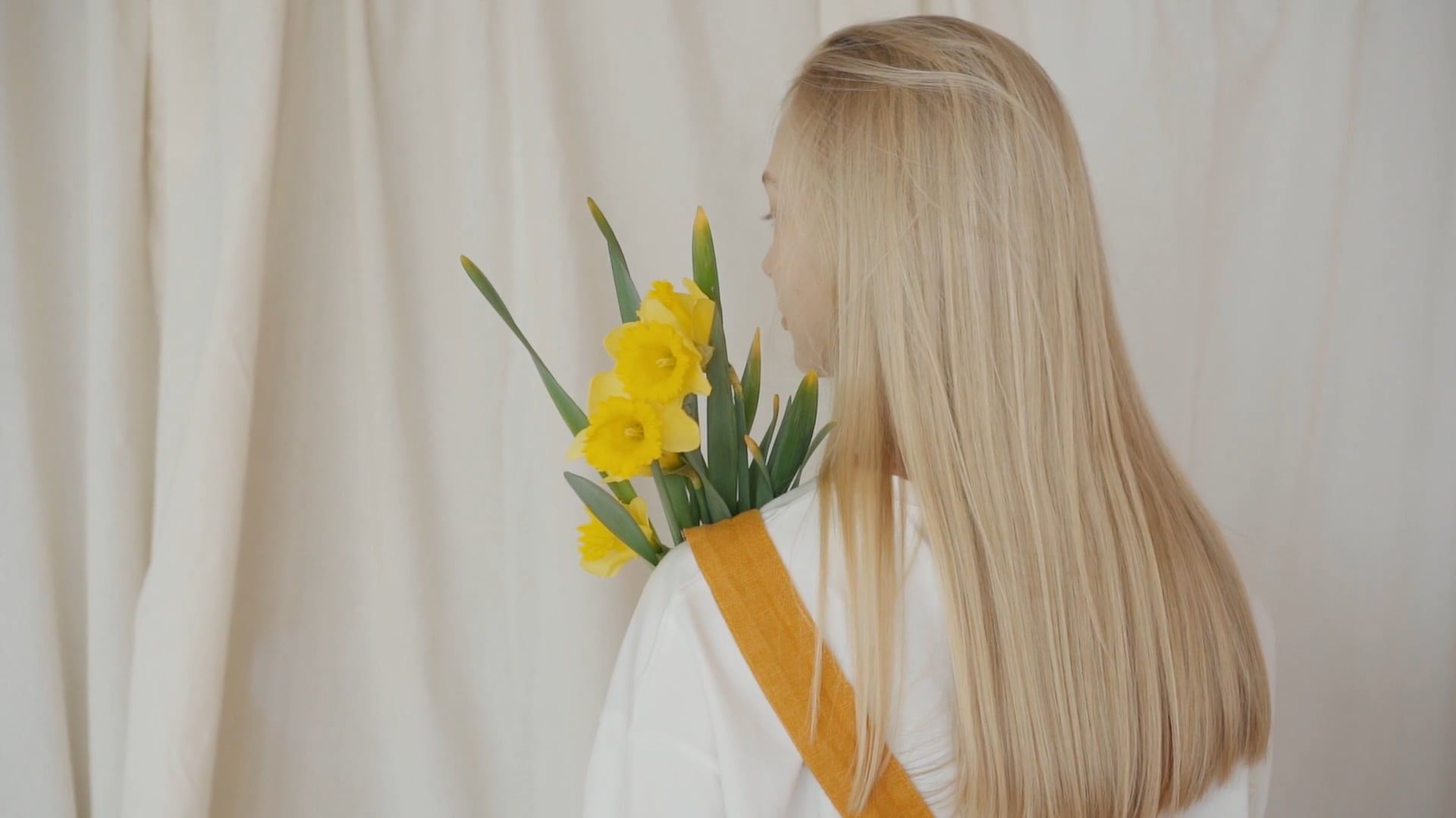 Mujer rubia de espaldas, con el cabello largo y liso, sosteniendo narcisos frente a una cortina crema